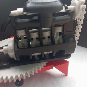 Puede incluir: Un modelo de motor de combustión interna impreso en 3D con un bloque de motor negro, cuatro pistones gris claro, una cadena de distribución blanca y un componente circular negro grande en la parte superior. Montado sobre una base de engranajes roja y blanca.