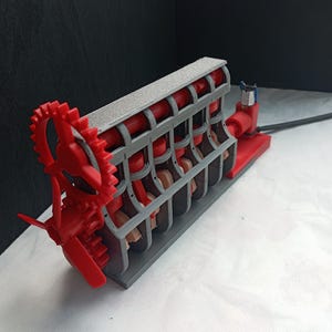 May include: A mechanical engine model featuring a grey frame with red internal components, including pistons and a gear assembly at the front. The structure is mounted on a rectangular base and includes a black cable extending from the rear section.