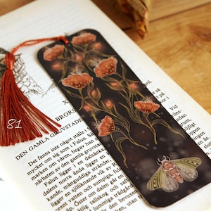 May include: A decorative bookmark with a dark background, orange floral illustrations, and a moth design. It has a red tassel. The number "81" is visible on the tassel. The bookmark is placed on an open book with text.