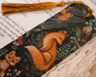 May include: A rectangular bookmark featuring a dark forest background with two orange foxes, small red mushrooms, and green foliage. The bookmark has a hole at the top with a gold-colored tassel attached, resting against a page of printed text.