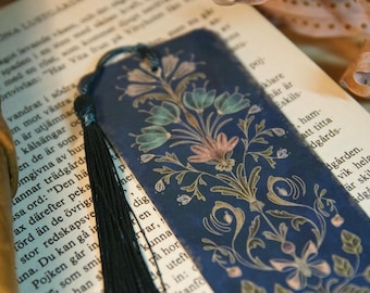May include: A navy blue bookmark with a floral design in shades of green, pink, and gold. The bookmark has a black tassel and is resting on the pages of a book.