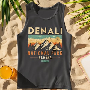 May include: Dark gray tank top with a vintage-style graphic featuring the text "DENALI" above a mountain scene with the words "NATIONAL PARK ALASKA" and "EST. 1917". The top is displayed on a sandy surface with palm leaves and sunglasses.