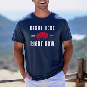 May include: Navy blue t-shirt with the text "RIGHT HERE RIGHT NOW" in white. A red bison graphic is centered between the years 2025 and 2026. The shirt is a crew neck style.
