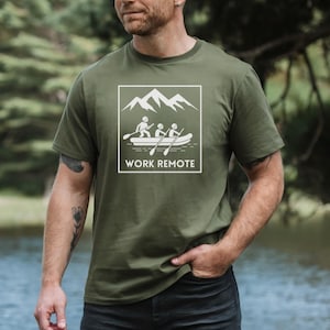 May include: Olive green t-shirt with a white graphic of a raft with three people and the text "WORK REMOTE" inside a white square. The shirt is short-sleeved and made of a soft material.