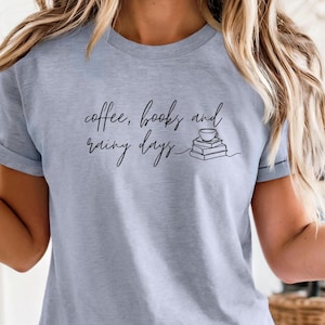 K&ouml;nnte beinhalten: Hellblaues T-Shirt mit dem Text "coffee, books and rainy days" in handschriftlicher Schrift. Das Design beinhaltet eine kleine Illustration einer Kaffeetasse auf einem Stapel B&uuml;cher. Es ist ein l&auml;ssiges Kleidungsst&uuml;ck f&uuml;r den Alltag.