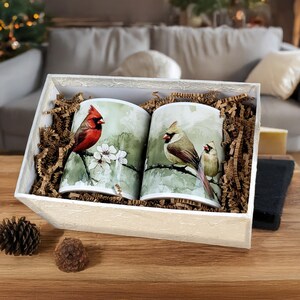 May include: Two ceramic mugs with cardinal bird illustrations are presented in a white gift box with brown paper filler. One mug features a red cardinal on a flowering branch. The other mug shows a female cardinal and a juvenile bird.
