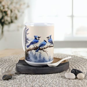 May include: A white ceramic mug featuring a watercolor painting of three blue jays perched on a tree branch. The background depicts a forest scene in shades of blue and green. The mug is on a wooden coaster.