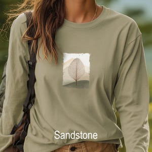 May include: Sandstone-colored long-sleeve shirt with a rectangular graphic of a leaf and mountain landscape. The leaf is a detailed, light brown skeleton leaf. The word "Sandstone" is printed at the bottom of the shirt.