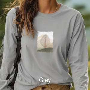 May include: Grey long-sleeve shirt with a nature-themed graphic. The graphic features a detailed leaf design set against a backdrop of mountains and earth tones. The word "Grey" is printed at the bottom of the shirt.