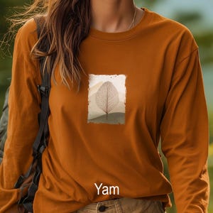 May include: A long-sleeved, burnt orange shirt with a rectangular graphic of a leaf skeleton against a mountain landscape. The word "Yam" is printed at the bottom of the shirt.