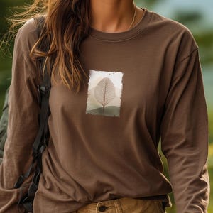 Pode incluir: Uma camisa marrom de manga comprida com um gráfico retangular de uma folha e uma paisagem montanhosa. A camisa é de cor marrom lisa e tem gola redonda.