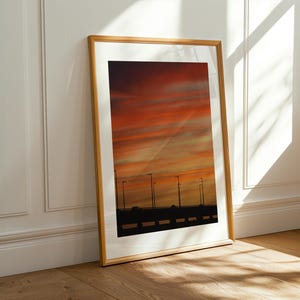 Puede incluir: Una impresión enmarcada de una vibrante puesta de sol con tonos naranja y rojo. La obra de arte presenta una silueta de un puente con farolas. El marco es de color madera clara, y la impresión está apoyada contra una pared blanca y un suelo de madera.