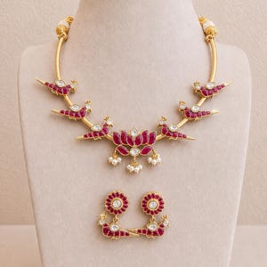 Puede incluir: Un conjunto de collar y pendientes a juego en tono dorado. El collar presenta un colgante de flor de loto y detalles en forma de pájaro con piedras preciosas rojas. Los pendientes tienen un diseño floral con una gema central. Joyas expuestas en un maniquí.