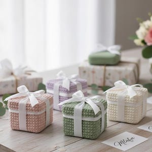 May include: A collection of small, square crochet gift boxes in pastel colors: pink, green, and cream. Each box is tied with a white satin ribbon. The boxes are arranged on a light wooden surface.