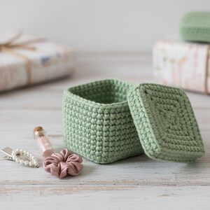 May include: A light green, square crochet box with a matching lid sits open, revealing its interior. A small pink scrunchie, a pearl hair clip, and a tiny glass bottle with pink contents are next to the box. The box is handmade and appears to be a storage container.