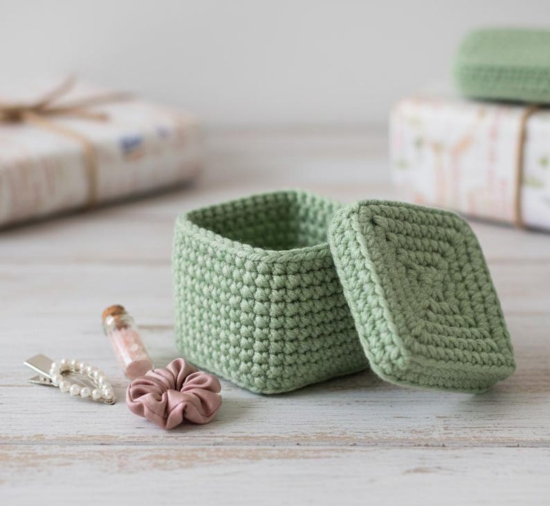 May include: A light green, square crochet box with a matching lid sits open, revealing its interior. A small pink scrunchie, a pearl hair clip, and a tiny glass bottle with pink contents are next to the box. The box is handmade and appears to be a storage container.