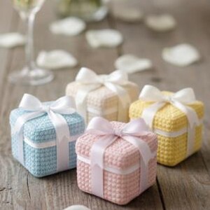 May include: Four small, square gift boxes in pastel colors: blue, pink, cream, and yellow. Each box is tied with a white satin ribbon bow. The boxes sit on a rustic wooden surface, with a glass of champagne and flower petals in the background.