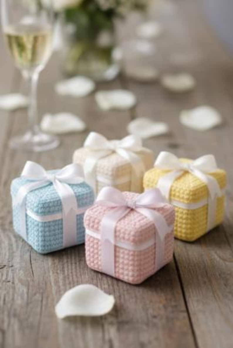 May include: Four small, square gift boxes in pastel colors: blue, pink, cream, and yellow. Each box is tied with a white satin ribbon bow. The boxes sit on a rustic wooden surface, with a glass of champagne and flower petals in the background.