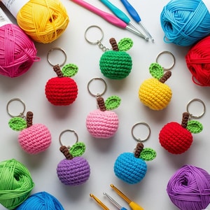 Könnte beinhalten: Eine Sammlung handgefertigter Apfel-Schlüsselanhänger aus Häkelarbeit in verschiedenen Farben, darunter Rot, Grün, Gelb, Rosa, Blau und Lila. Jeder Schlüsselanhänger hat einen braunen Stiel und grüne Blätter. Bunte Wolle und Häkelnadeln sind ebenfalls zu sehen.