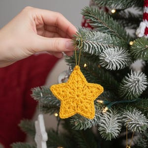 Puede incluir: Adorno navideño de estrella amarilla de ganchillo con hilo dorado, sostenido cerca de un árbol de Navidad decorado. La estrella está hecha de hilo y tiene una superficie texturizada. El árbol tiene agujas verdes y está decorado con luces.