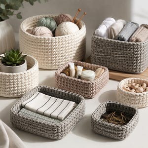 May include: A collection of handmade crochet baskets in various sizes and colors. Some baskets contain yarn, toiletries, and a succulent plant. The baskets are in shades of cream, gray, and pink, offering versatile storage solutions.