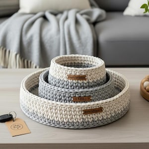 May include: Three handmade crochet baskets in a nested set. The baskets are in shades of white and grey, with a round design. Each basket has a small, rectangular label with text on it.