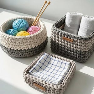 May include: A set of three handmade crochet baskets in neutral tones. The round basket holds yarn and knitting needles. The rectangular basket contains rolled white towels. The square basket holds a folded checkered cloth.