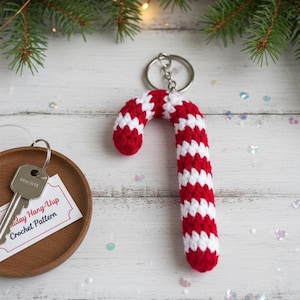 May include: A red and white crocheted candy cane keychain. The candy cane is in a classic striped pattern and attached to a silver keyring. A key and a small card are in the background.