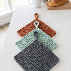 Puede incluir: Tres salvamanteles de ganchillo hechos a mano en tonos óxido, verde agua y gris carbón. Cada salvamanteles cuadrado tiene un lazo para colgar. Los salvamanteles están dispuestos en diagonal sobre una superficie blanca.