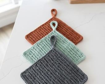 May include: Three handmade crochet pot holders in shades of rust, seafoam green, and charcoal gray. Each square pot holder has a loop for hanging. The pot holders are arranged diagonally on a white surface.