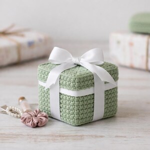 May include: A small, square, sage green crocheted gift box tied with a white satin ribbon and bow. The box is sitting on a light-colored wooden surface. Other wrapped gifts are in the background.