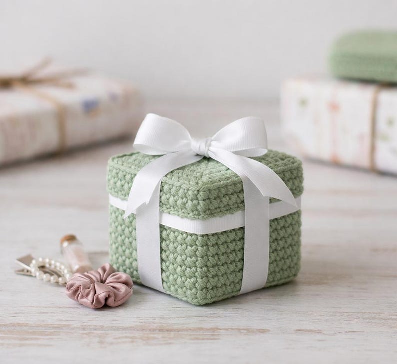 May include: A small, square, sage green crocheted gift box tied with a white satin ribbon and bow. The box is sitting on a light-colored wooden surface. Other wrapped gifts are in the background.