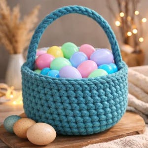 Op de afbeelding: Een turquoise gehaakte mand gevuld met pastelkleurige paaseieren. De mand heeft een gevlochten handvat en staat op een houten oppervlak. Verschillende gespikkelde eieren liggen voor de mand, wat een lentethema suggereert.
