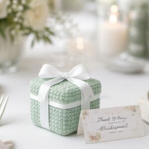 May include: A small, square, mint green crocheted gift box tied with a white satin ribbon. A floral-themed card with the words "Thank You for Being Bridesmaid!" is placed next to the box. The scene is set on a white surface with soft, blurred background elements.