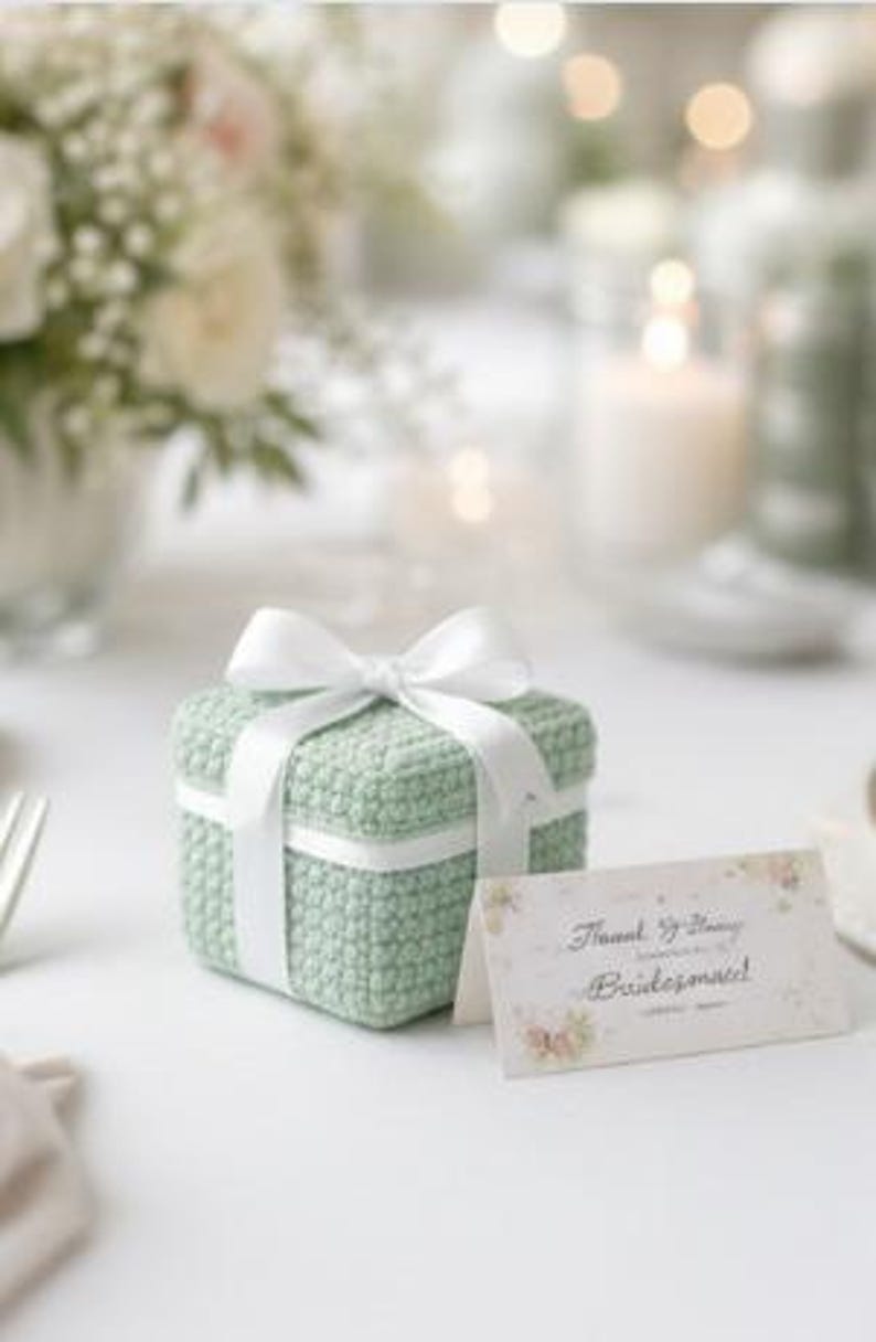 May include: A small, square, mint green crocheted gift box tied with a white satin ribbon. A floral-themed card with the words "Thank You for Being Bridesmaid!" is placed next to the box. The scene is set on a white surface with soft, blurred background elements.