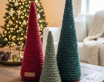 Könnte beinhalten: Drei gehäkelte Weihnachtsbäume in Rot, Grün und Grau stehen auf einem Holztisch. Die Bäume sind kegelförmig und haben ein strukturiertes Aussehen. Im Hintergrund befindet sich ein Weihnachtsbaum mit Lichtern.