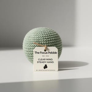 May include: A light green, crocheted sphere with a hang tag that reads "The Focus Pebble." The tag also includes the text "Clear Mind. Steady Hand." and the Hearthcraft Studio Co. logo. The sphere is approximately 7.6 cm in diameter.