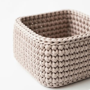 May include: A square, beige crochet basket. The basket is made of thick, woven yarn, with a textured surface. The basket's sides are straight, and the top edge is neatly finished. The neutral color and simple design make it suitable for various uses.