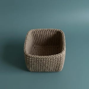 May include: A beige, square-shaped crochet basket. The basket has a rounded top and is made of thick, textured yarn. The basket is empty and sits against a teal background. This basket is ideal for storage.