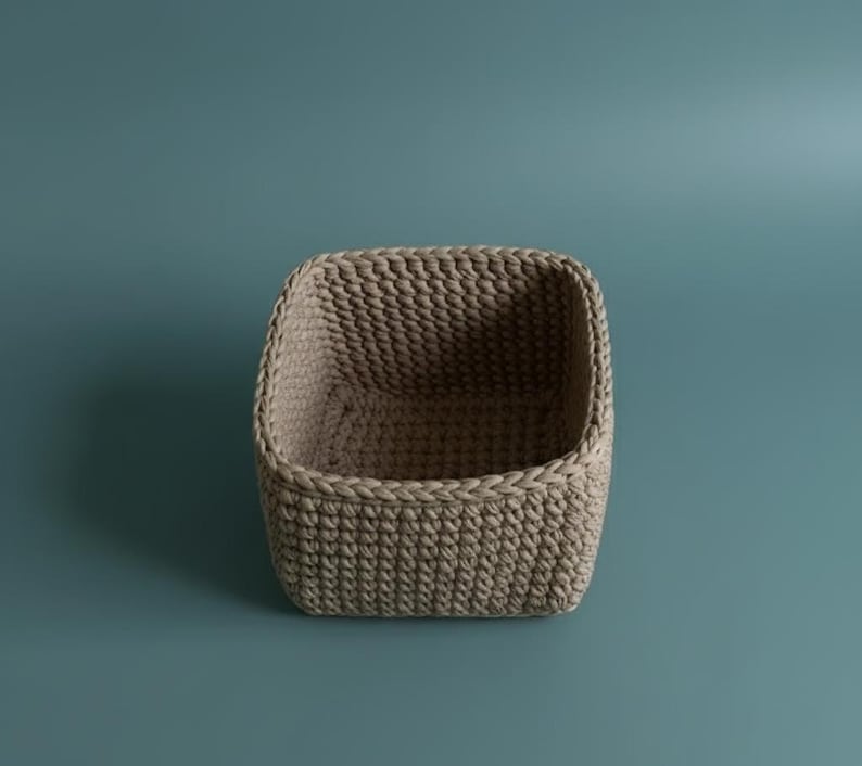 May include: A beige, square-shaped crochet basket. The basket has a rounded top and is made of thick, textured yarn. The basket is empty and sits against a teal background. This basket is ideal for storage.