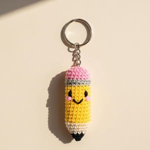 Könnte beinhalten: Ein handgefertigter Bleistift-Schlüsselanhänger aus Häkelarbeit mit einem lächelnden Gesicht. Der Bleistift hat einen gelben Körper, einen rosa Radiergummi, eine graue Zwinge und eine schwarze Spitze. Der Schlüsselanhänger ist an einem silbernen Metallring befestigt.