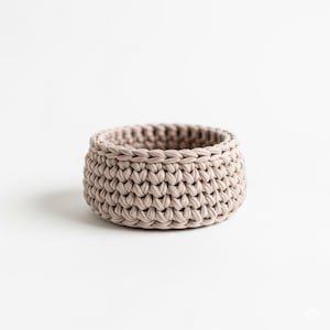 May include: A small, round, beige crochet basket. The basket is made of thick yarn and has a textured appearance. The basket is suitable for storing small items.