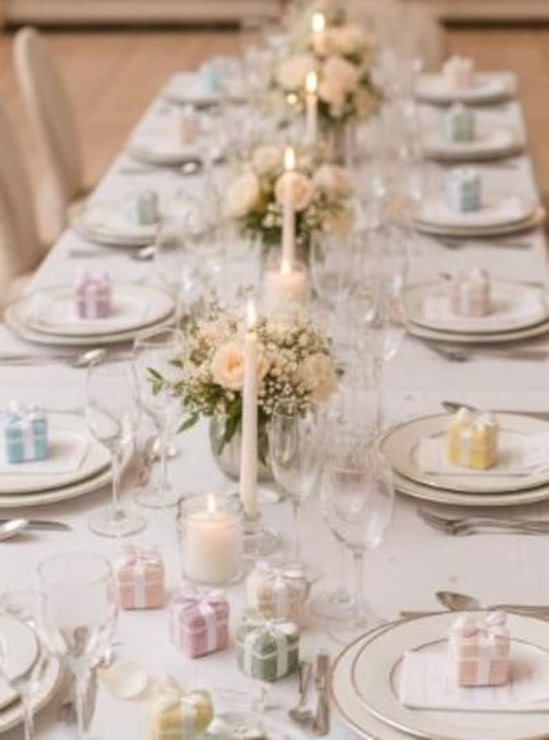 May include: Elegant table setting with white tablecloth, plates, and silverware. Centerpieces of roses and candles. Small gift boxes in pastel colors are placed at each setting. Wine glasses are arranged around the plates.