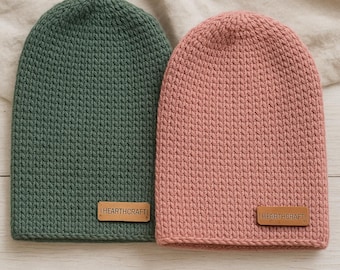 May include: Two knitted beanies, one in a muted green and the other in a dusty rose color. Each beanie has a rectangular leather patch with the text "HEARTHCRAFT" stitched on the front. The beanies are displayed on a light-colored, textured surface.