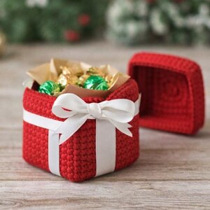 May include: A red crocheted gift box with a white ribbon and bow. The box is open, revealing wrapped candies inside. The lid is placed next to the box. The box is sitting on a wooden surface.