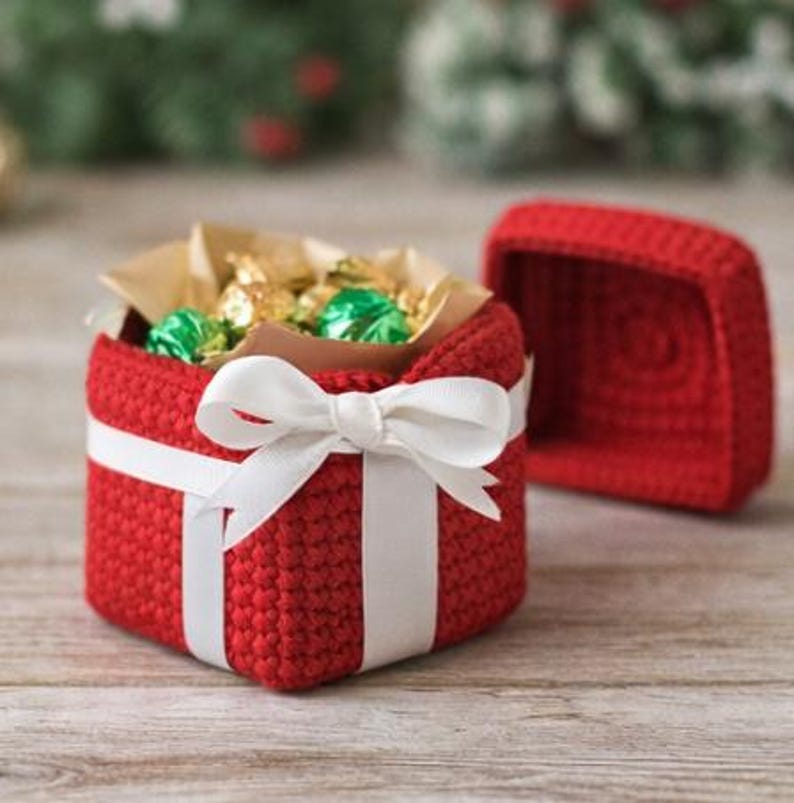 May include: A red crocheted gift box with a white ribbon and bow. The box is open, revealing wrapped candies inside. The lid is placed next to the box. The box is sitting on a wooden surface.
