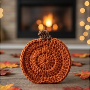 May include: A handmade, orange crochet pumpkin with a brown stem. The pumpkin is round and sits on a wooden surface, surrounded by fall leaves. The background features a fireplace with a warm glow, creating a cozy autumn scene.