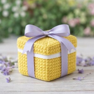 May include: A square, yellow crocheted box with a white ribbon and a lavender bow. The box is tied with a lavender ribbon that forms a bow on top. The background is blurred with greenery and flowers.