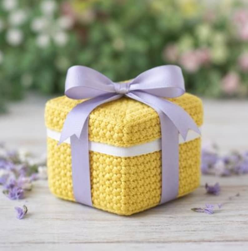 May include: A square, yellow crocheted box with a white ribbon and a lavender bow. The box is tied with a lavender ribbon that forms a bow on top. The background is blurred with greenery and flowers.