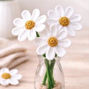 Puede incluir: Un jarrón de vidrio transparente contiene tres flores de margarita de ganchillo blancas con centros amarillos y tallos verdes. El jarrón descansa sobre pequeños guijarros. El texto "Classic Daisy Flower" y "Crochet Pattern" es visible.